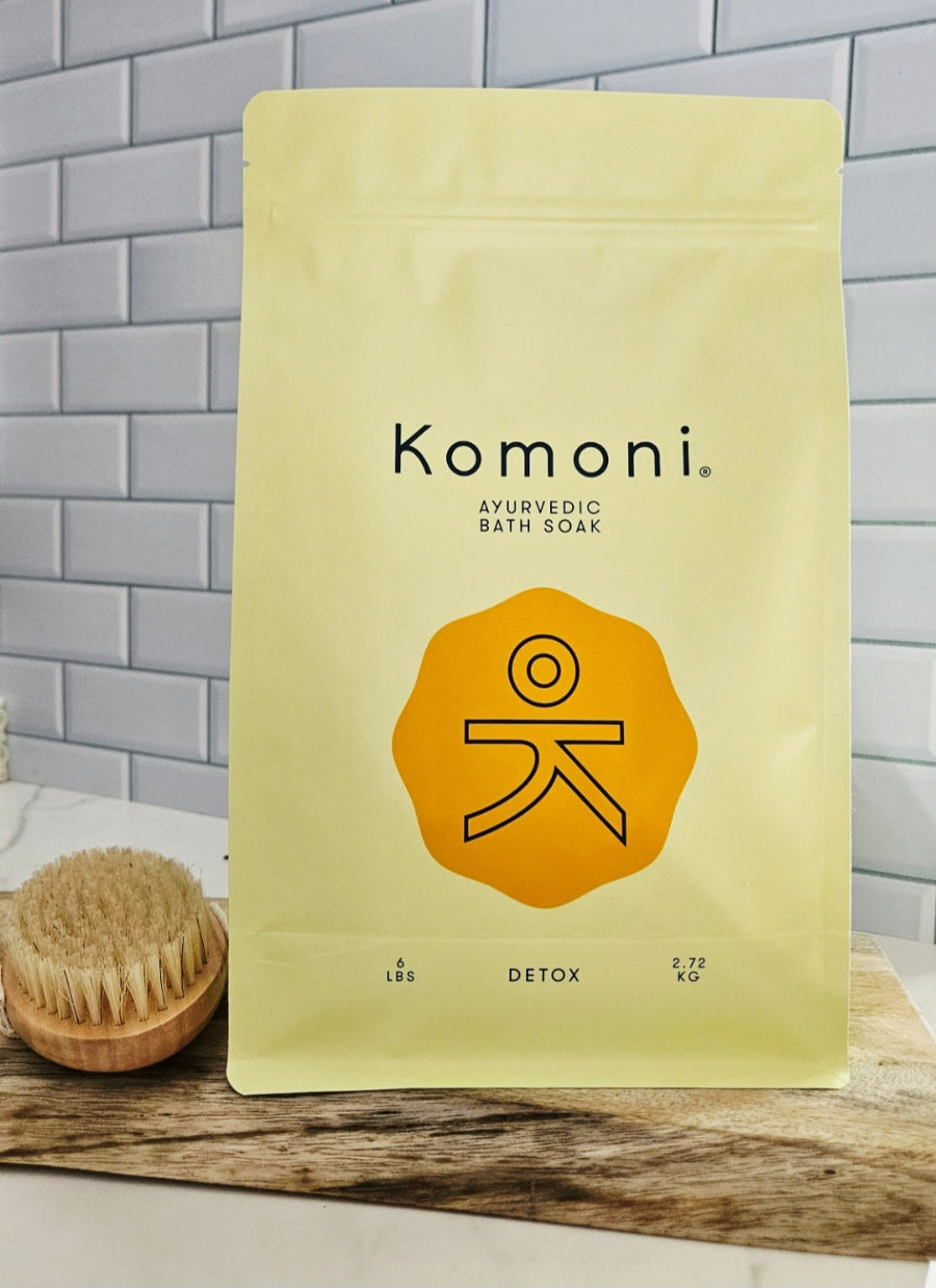 Komoni Ayurvedic Bath Soak 6 pound packaging with a wooden brush on a tiled bathroom floor.