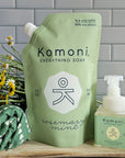 Komoni Everything Soap refill packaging with a smaller bottle and flowers on a wooden surface.