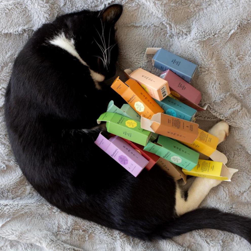 A black cat with white patches shown curled up with all the colorful boxes of Komoni's pure essential oils.