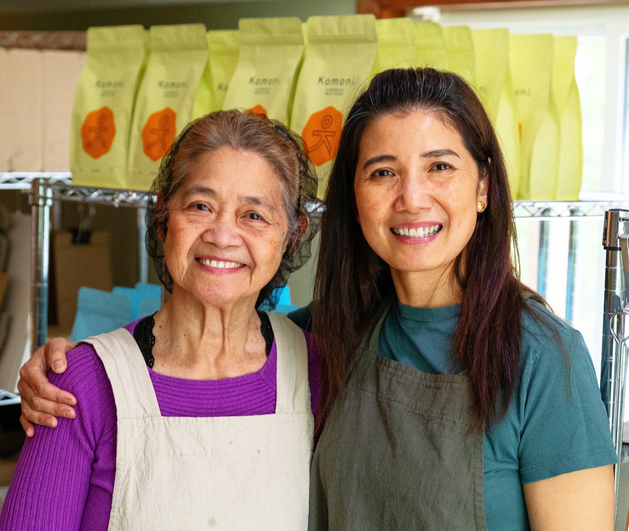 The two women that own Komoni standing together with shelves of yellow and blue bags of Bath Soak in the background