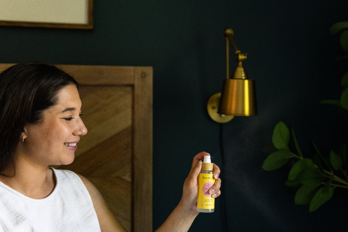 Woman spraying a yellow bottle of LAURENTHA Aromamist with a dark wall and plant in the background