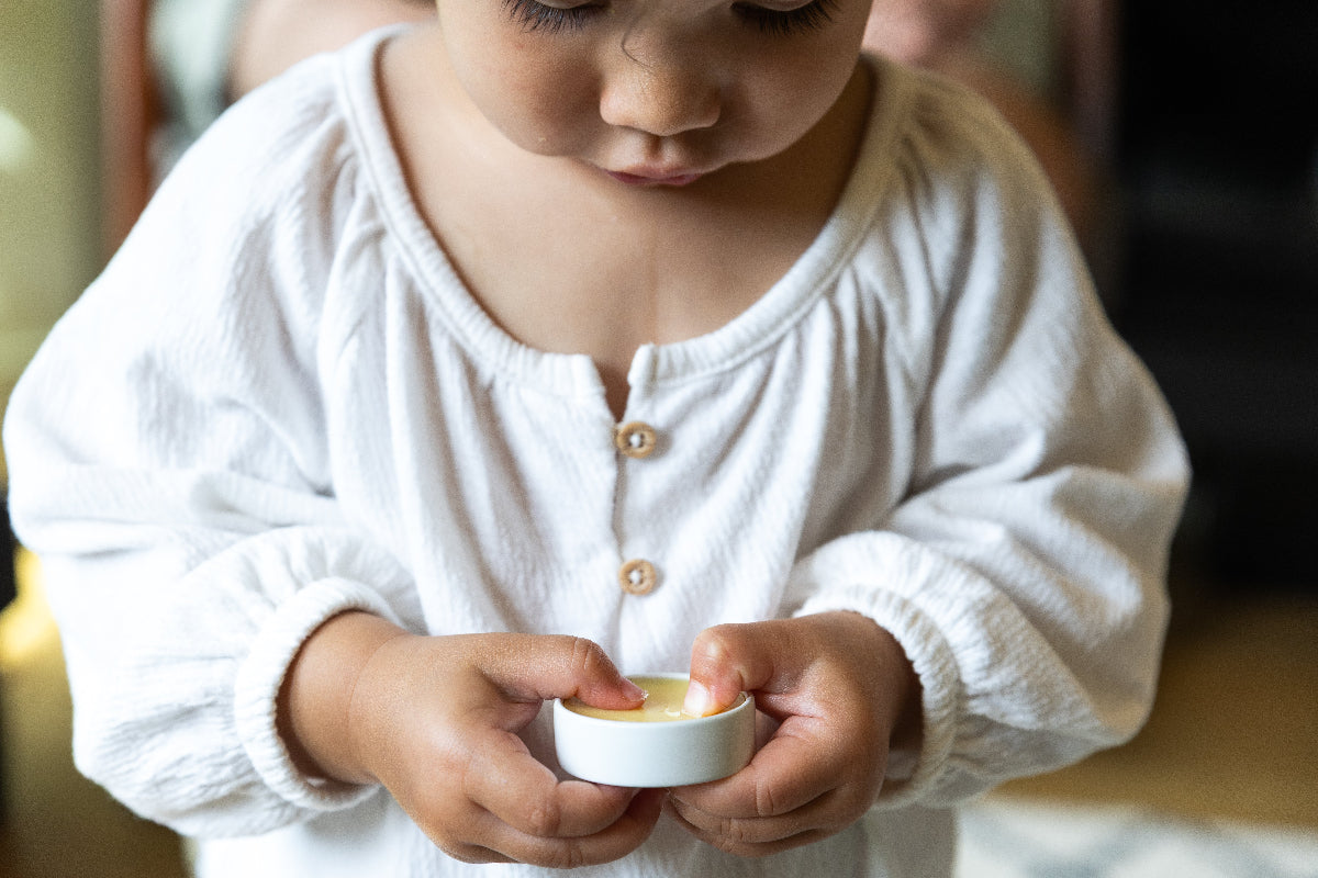 Child holding a small white open tin showing the light yellow Everything Salve inside, wearing a white long-sleeve shirt.