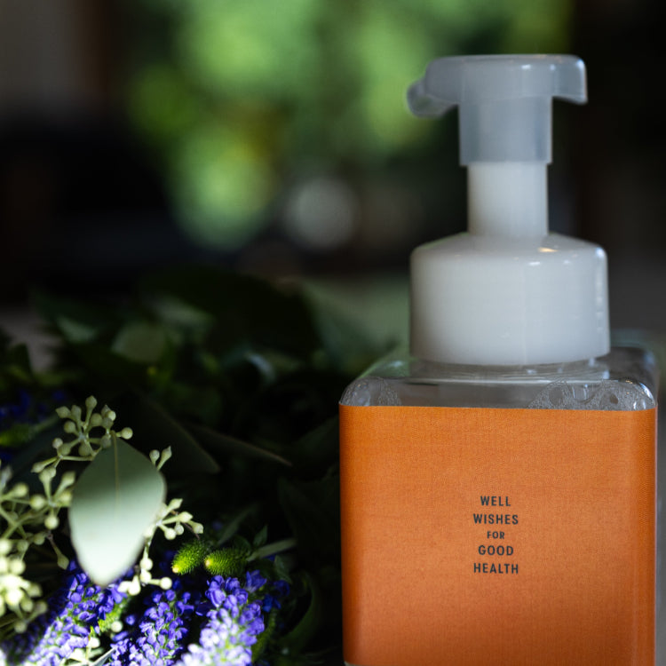 Foaming everything hand soap dispenser with 'Well Wishes for Good Health' text, surrounded by greenery.