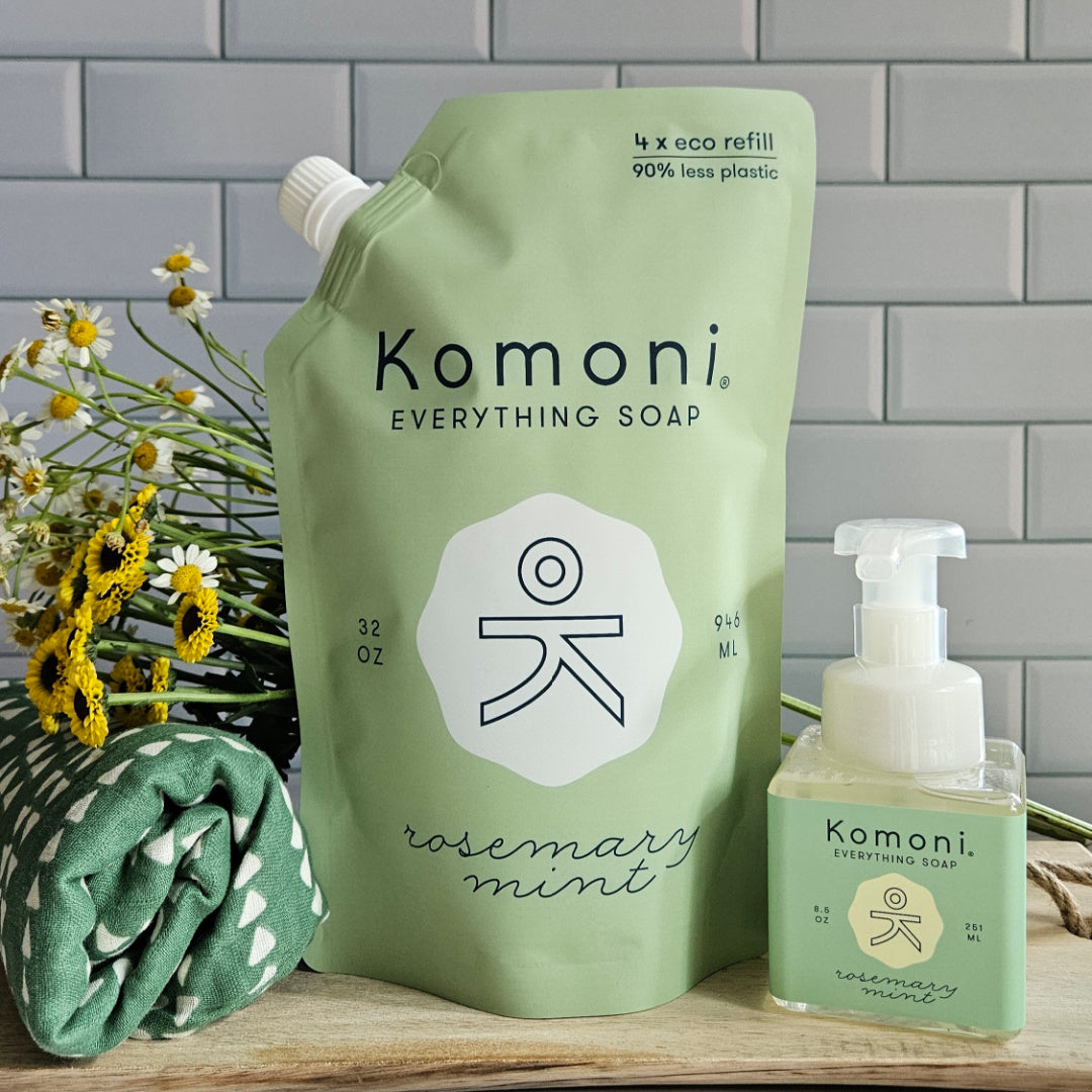 Komoni Everything Soap refill packaging with a smaller bottle and flowers on a wooden surface.