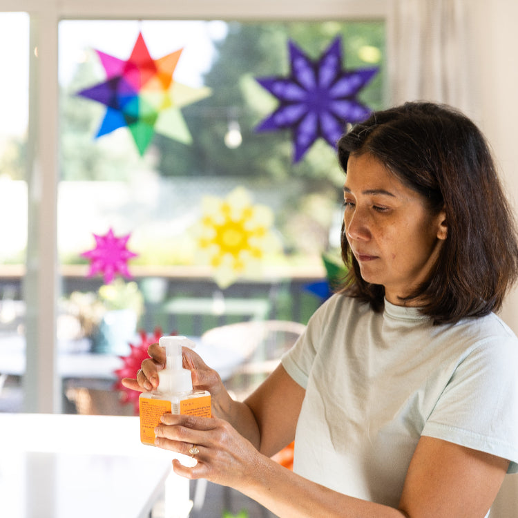 Komoni founder holding an orange colored dispenser her Everything Soap  with colorful decorations in the background.