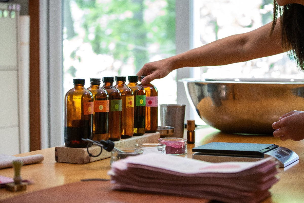 The making of my bath soak with small bowls of botanicals and bottles of my signature essential oil blends on a table with my hand reaching towards them, in a bright room.