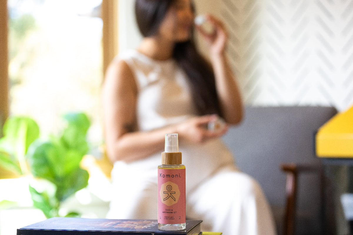 A bottle of aromamist body-room-linen spray with a blurred indoor background showing a woman smelling from a tin of everything salve. 