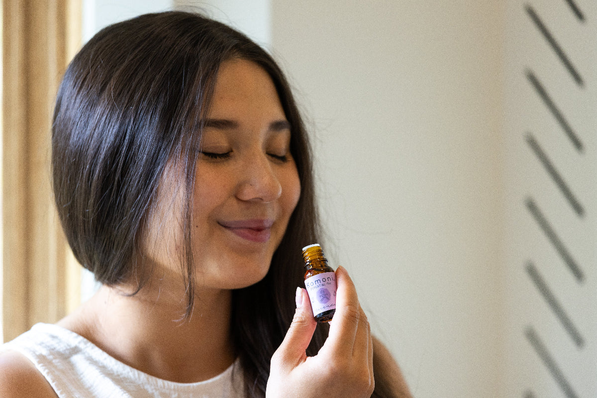 Woman holding and smelling a small, open vial of pure essential with a purple label against a neutral background