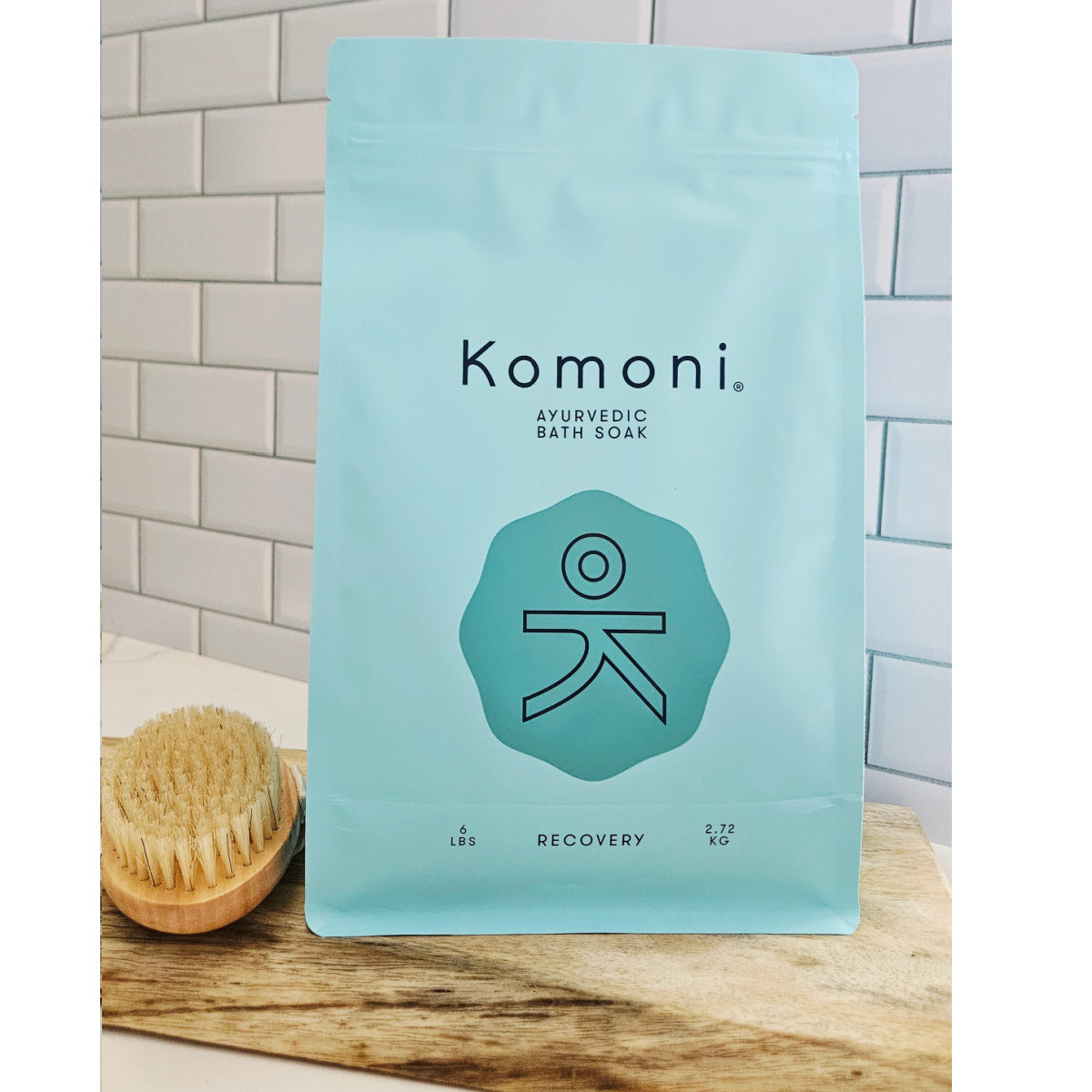 Komoni RECOVERY Ayurvedic Bath Soak 6 pound package on a wooden surface with a brush in the background.