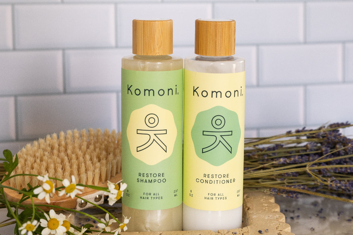 Two Komoni shampoo and conditioner bottles with bamboo caps on a white tiled background.